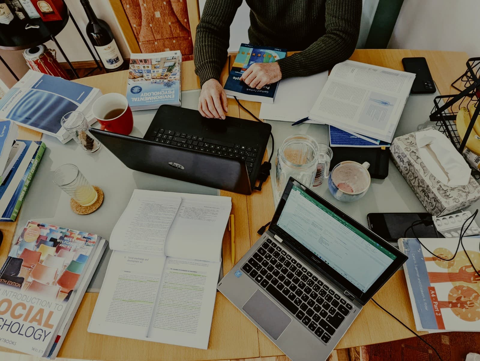 Person lernt an einem Tisch mit zwei Laptops, vielen Fachbüchern, Notizen und verschiedenen Getränken.