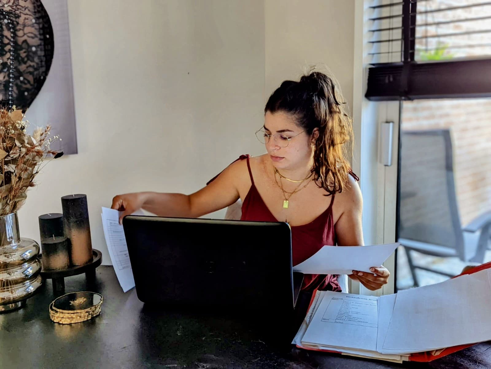 Frau mit Brille arbeitet konzentriert am Laptop und sichtet dabei verschiedene Dokumente am Schreibtisch.