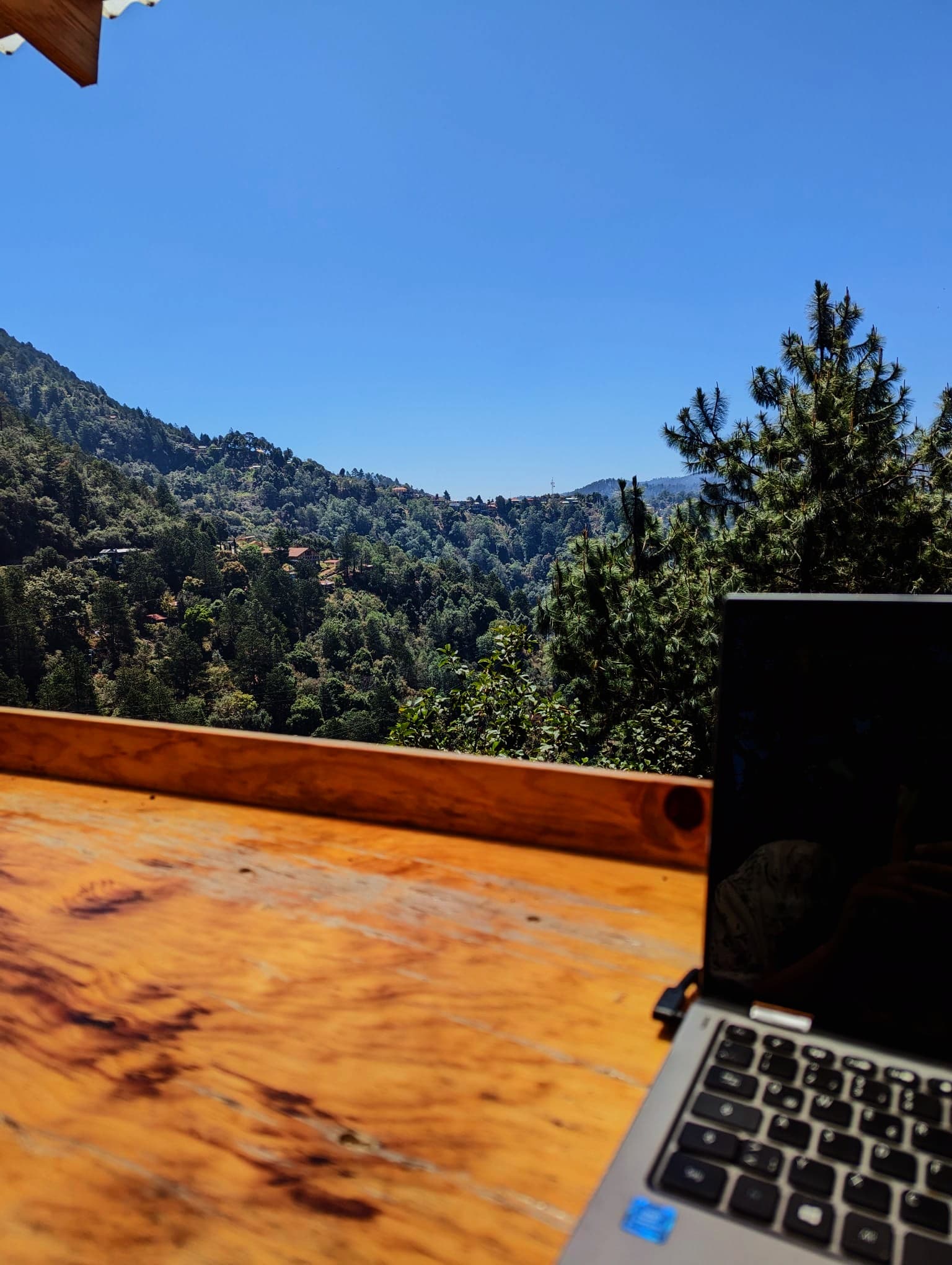 Laptop auf Holztisch vor einer weiten Aussicht auf bewaldete Berge und strahlend blauen Himmel.