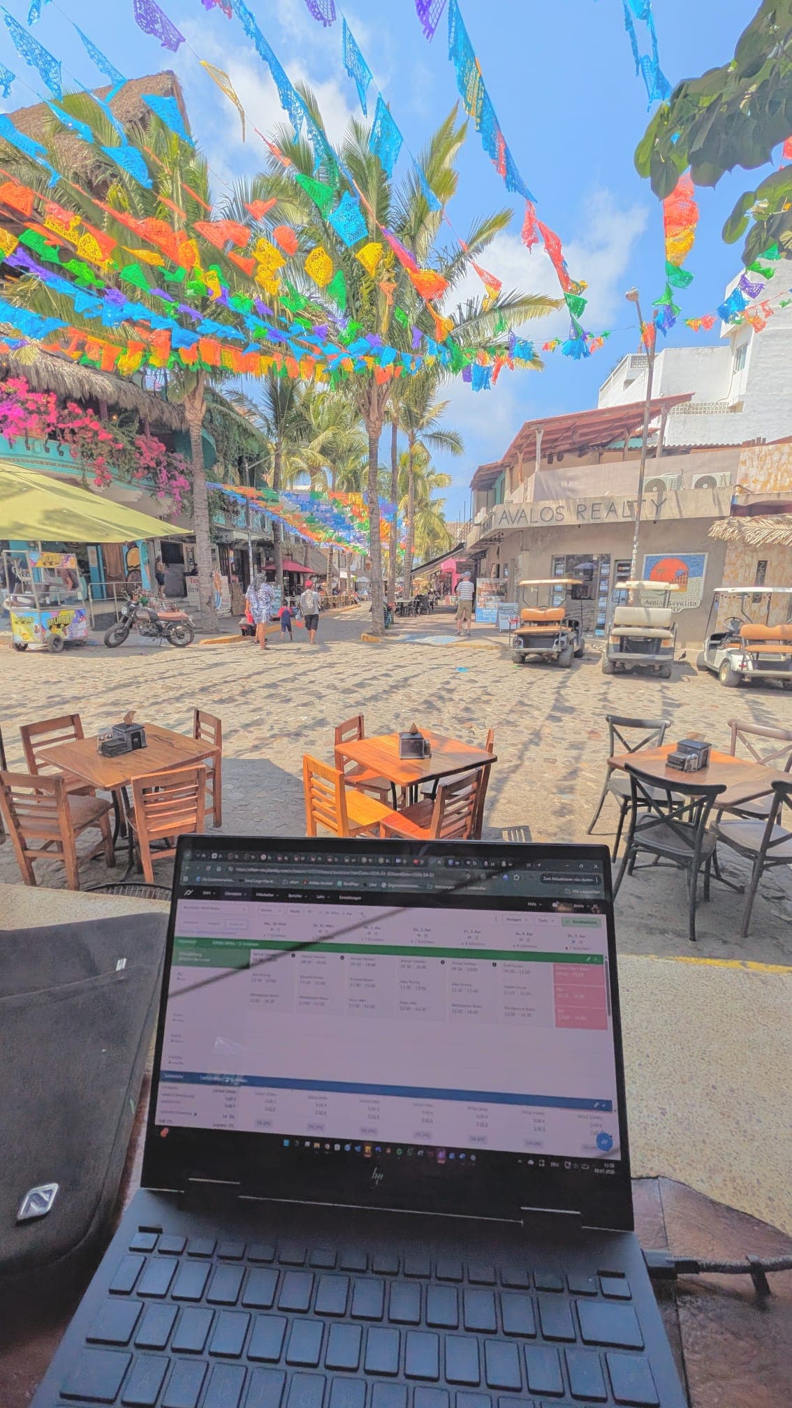 Laptop auf einem Cafétisch mit Blick auf eine sonnige Straße mit bunten mexikanischen Wimpelketten.