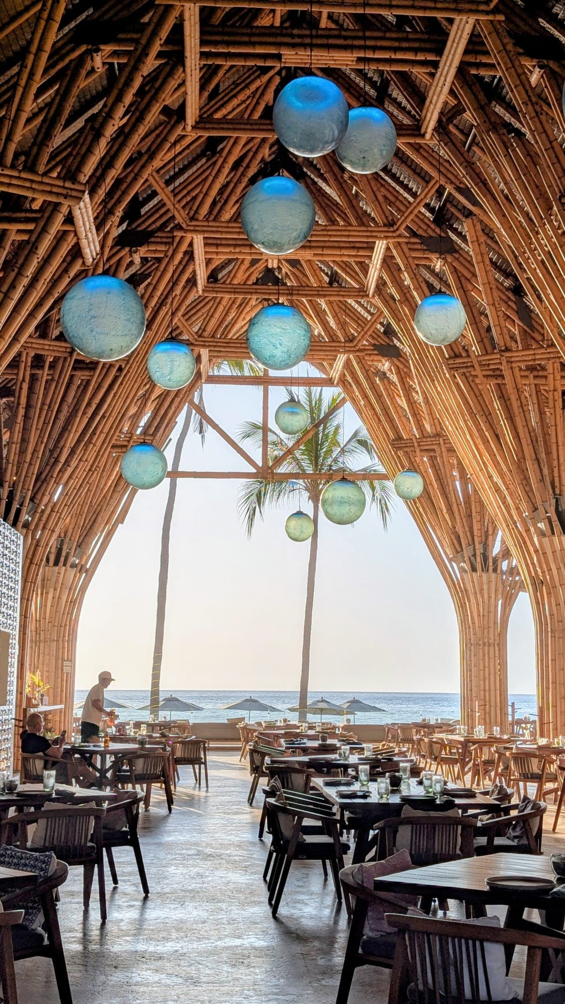 Hohes Bambusgewölbe in einem Restaurant mit blauen Hängeleuchten und Blick auf Strand und Meer.